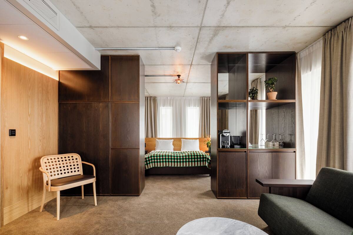 A compact hotel room with a bed, a seating area, a small table, and a built-in bookshelf.