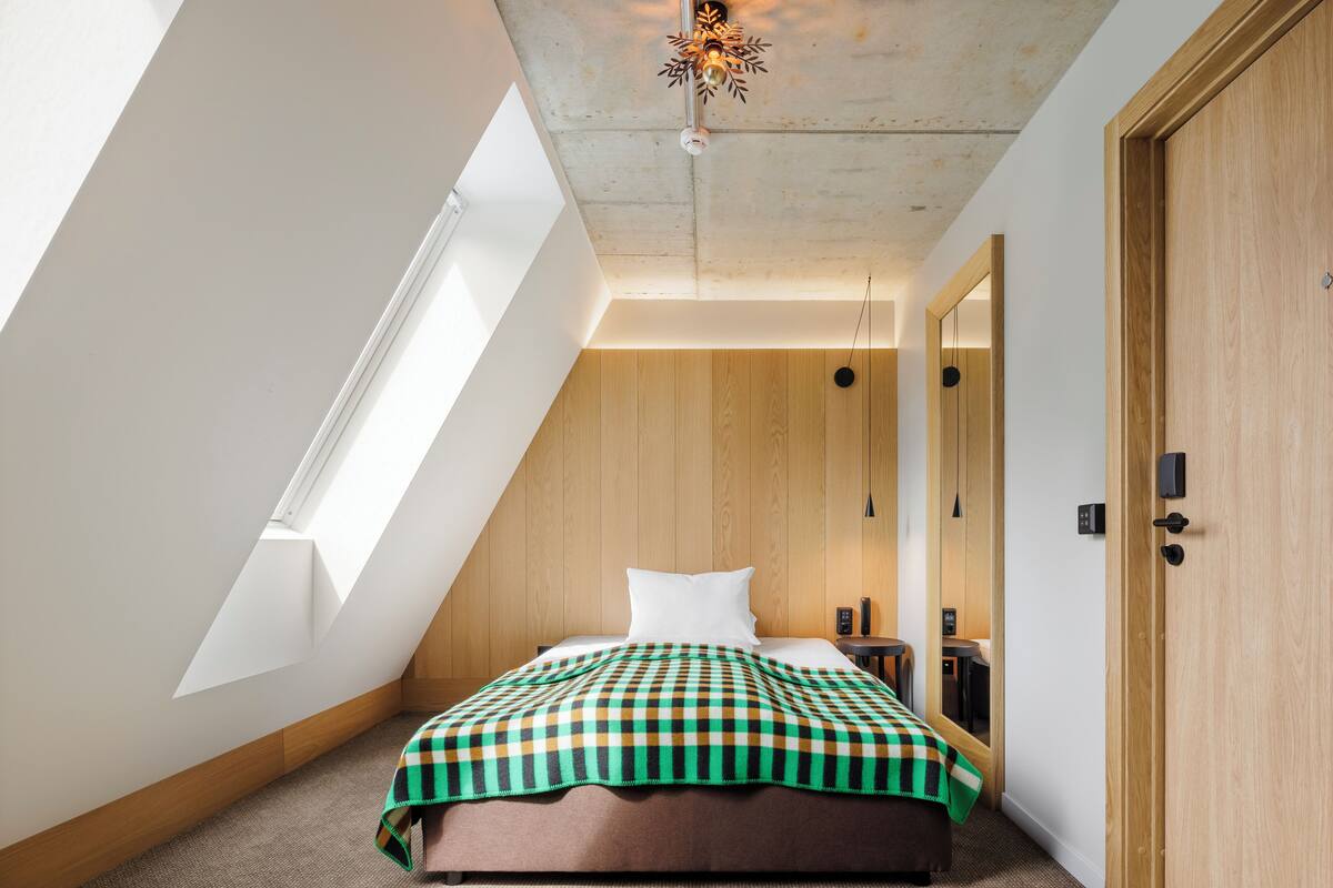 A bedroom with a sloped ceiling, a skylight, a bed with a checkered blanket, a bedside table, and a wooden wardrobe.