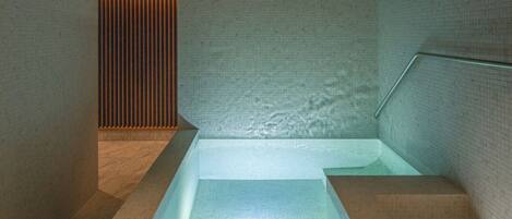 Un moderno espacio de spa con una bañera de hidromasaje rectangular, paredes alicatadas y un panel de madera en el fondo.