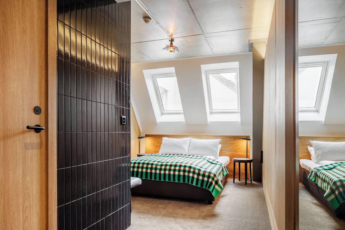 A bedroom with a sloped ceiling, skylights, a bed with a checkered bedspread, a bedside table, and a chair.