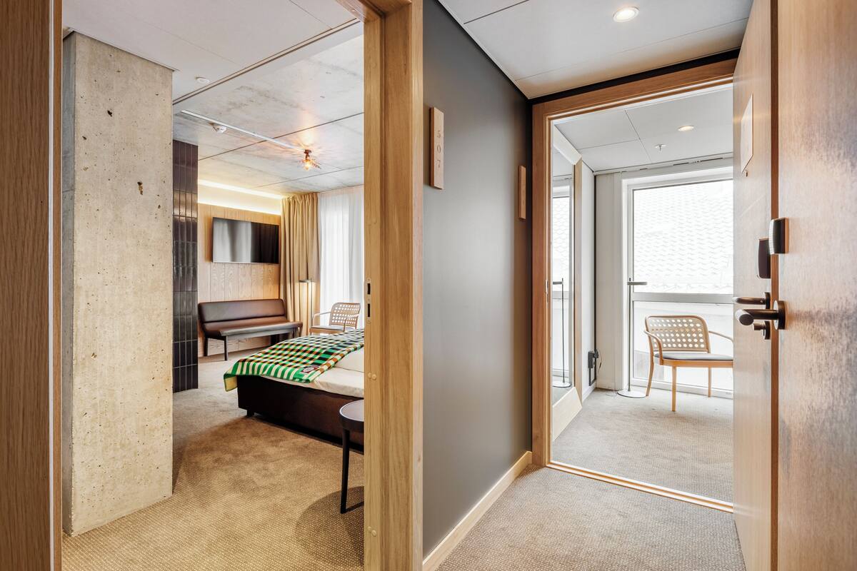A modern hotel room with a bed, a bench, a small table, and a chair. There is a door leading to another room and a window with a view.