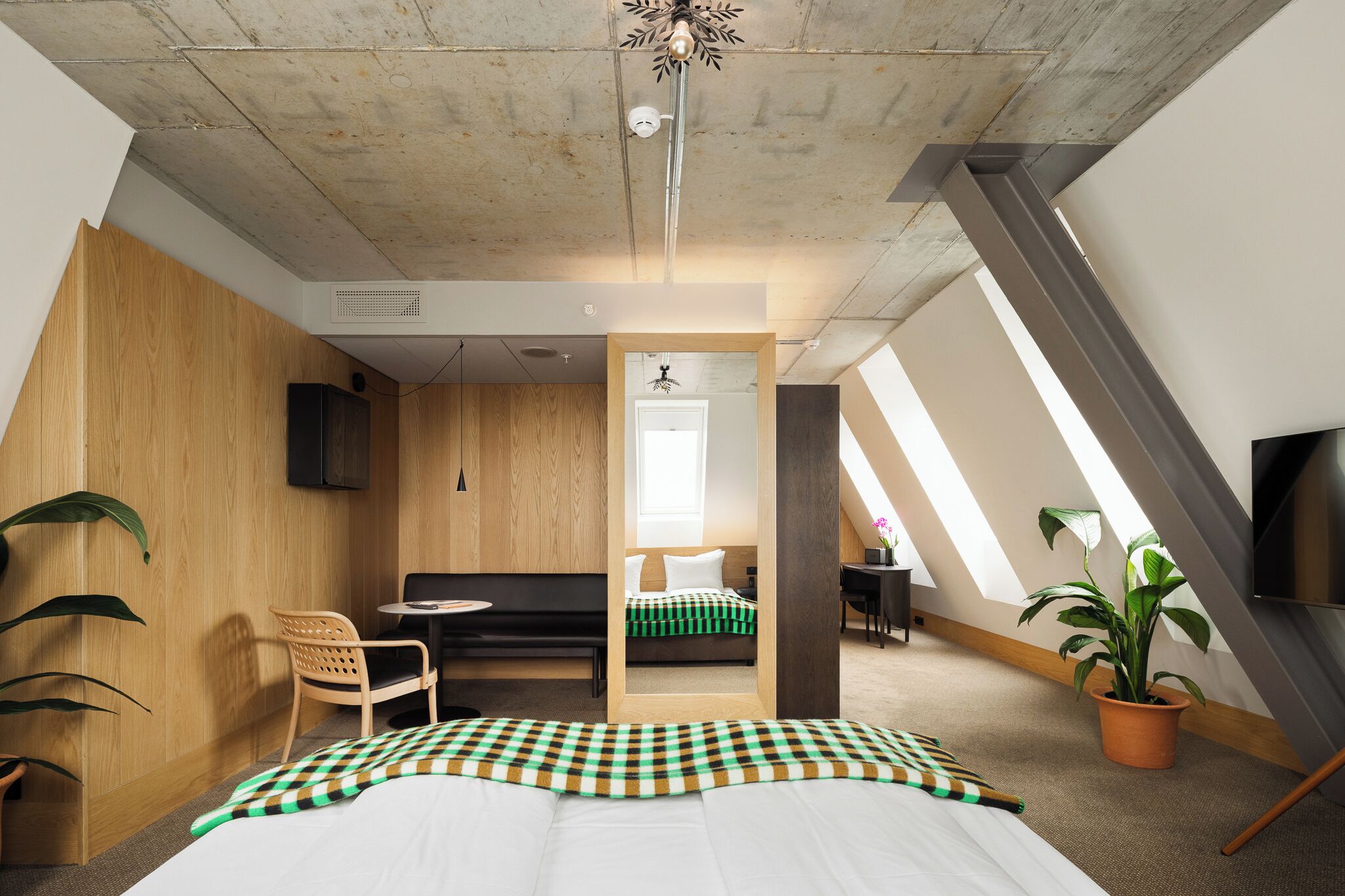 Une chambre moderne et compacte avec un plafond en pente, un lit recouvert d’une couverture à carreaux, un bureau avec une chaise, une télévision et une plante en pot.