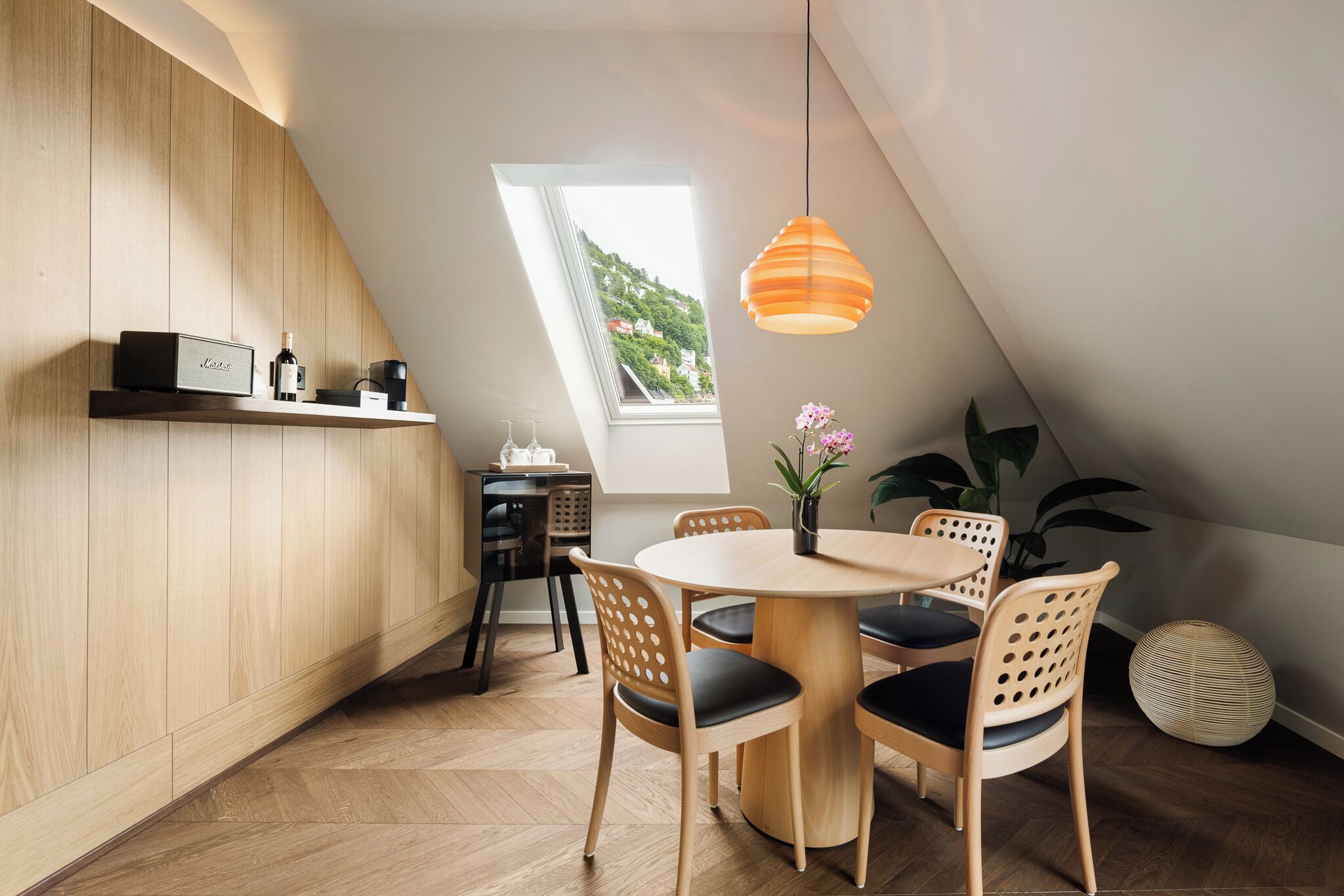 Ett modernt kök med träinredning, en matplats med ett runt bord och stolar samt ett stort fönster med utsikt över grönområden.