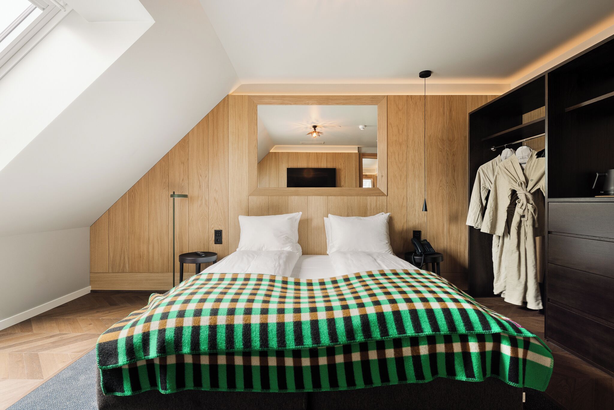 Un dormitorio con techo inclinado, una cama con ropa blanca, un plaid de cuadros, una mesita de noche y un armario.