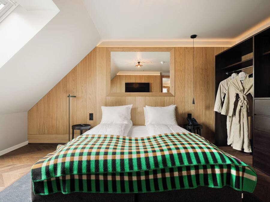 Un dormitorio con techo inclinado, una cama con ropa blanca, un plaid de cuadros, una mesita de noche y un armario.