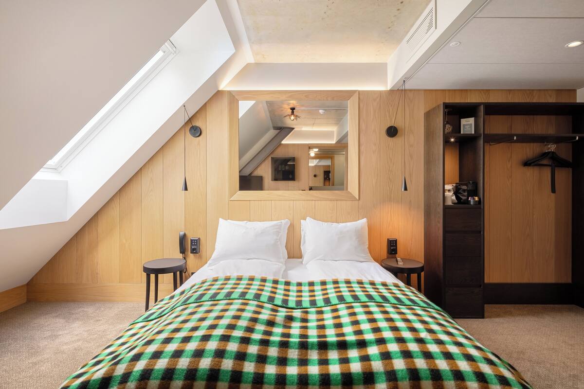 A bedroom with a sloped ceiling, a bed with a checkered blanket, a wooden wardrobe, and a mirror on the wall.