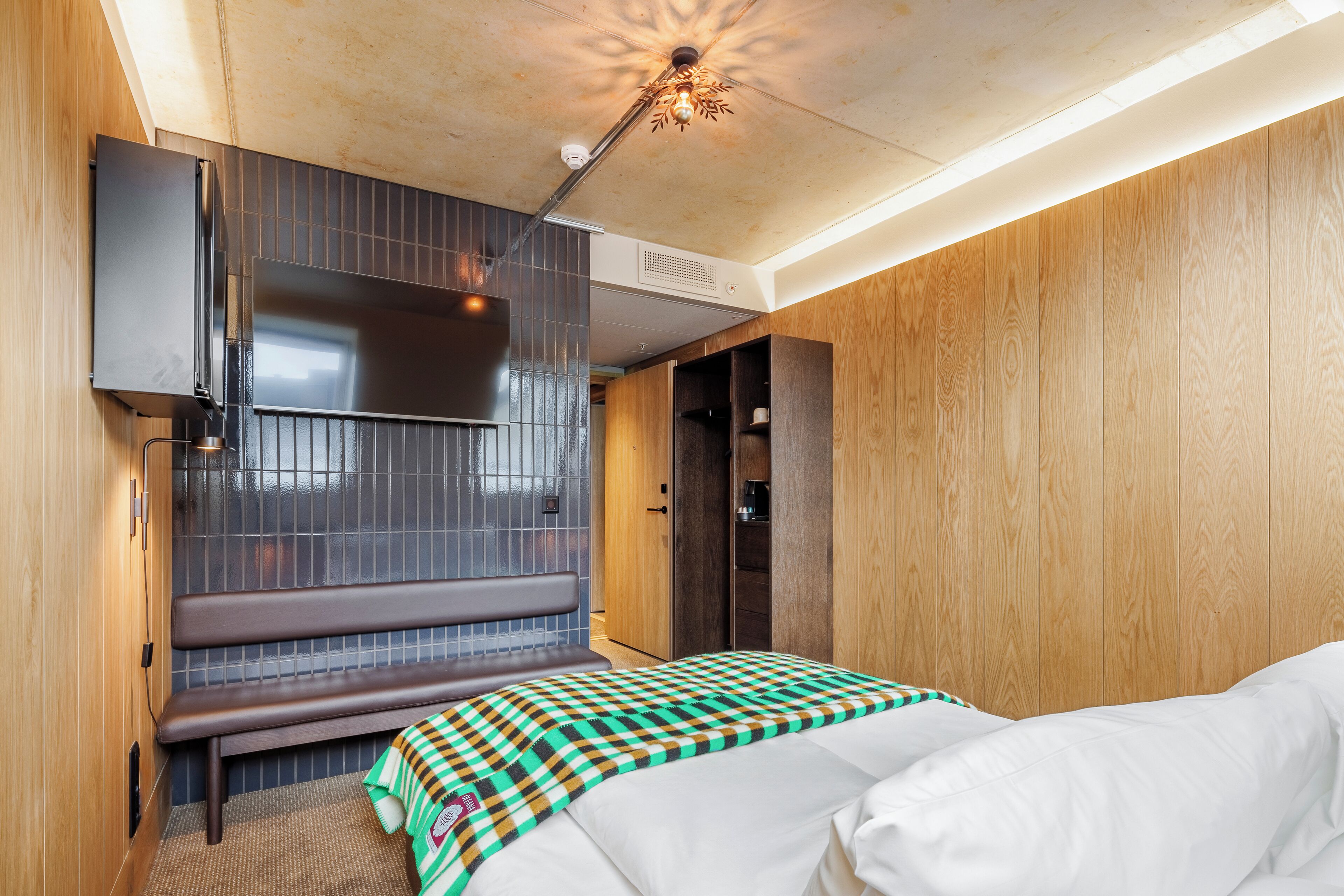 Quarto de hotel moderno com parede de madeira, um banco, uma cama com manta xadrez e luminária no teto.