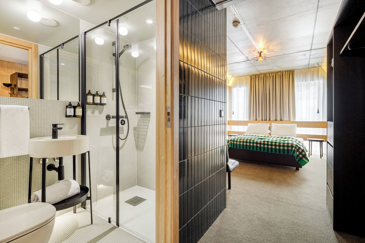 A modern hotel room with a glass-enclosed shower, a bedside table, a bed with a green checkered bedspread, and a large window with curtains.