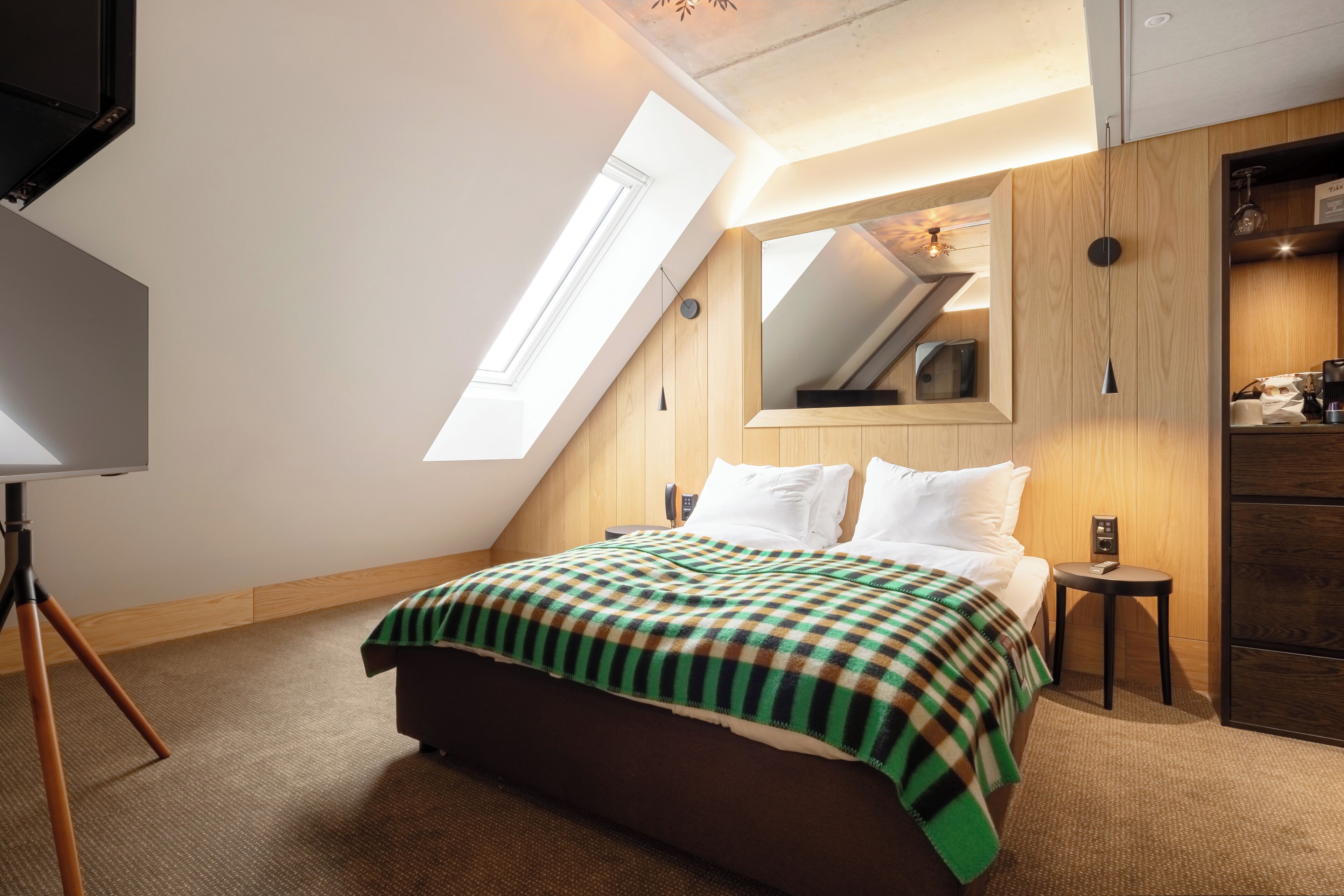 A bedroom with a skylight, a bed with a plaid blanket, a small table, and a wardrobe.