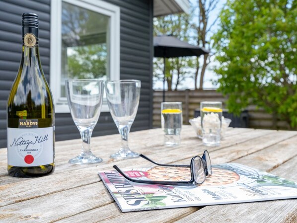 A bottle of Nottage Hill Chardonnay, two wine glasses, a pair of glasses, and a magazine on a wooden table outdoors.