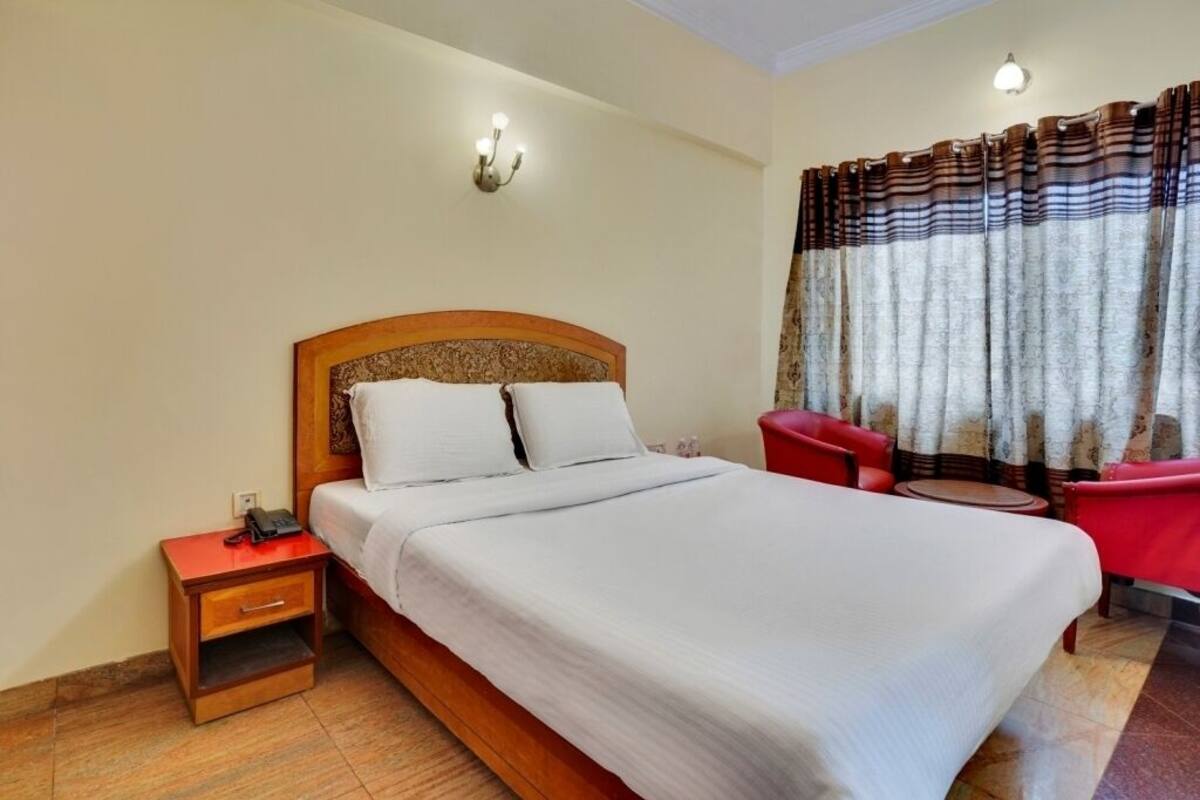 A neatly made bed with white linens, a wooden nightstand with a phone, and a red chair by a window with curtains.
