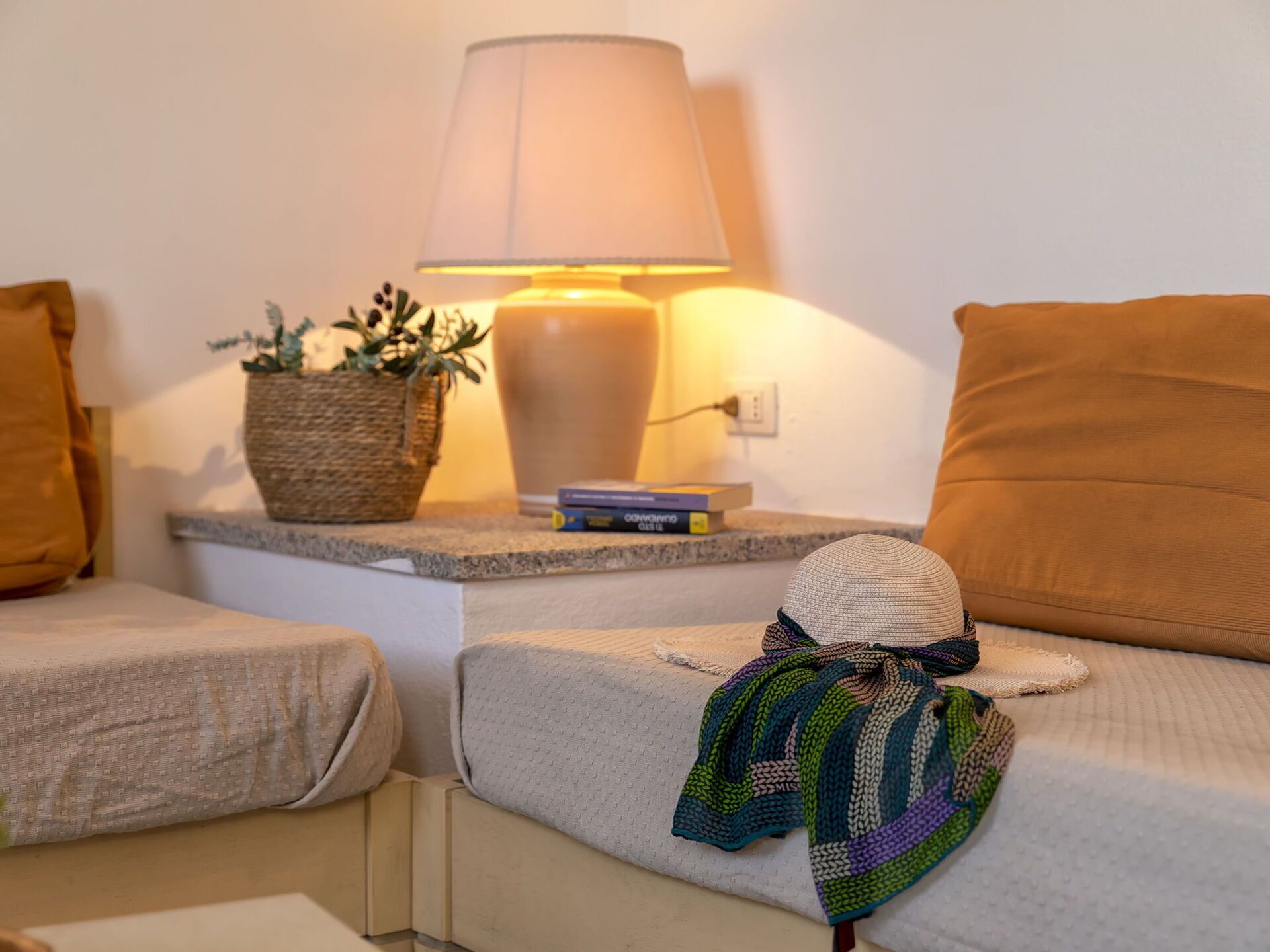 Habitación de hotel ordenada con una cama, una mesita de noche con lámpara, una planta pequeña en maceta y una pila de libros.