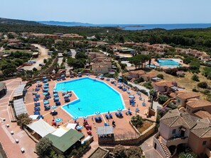 A resort with a central swimming pool surrounded by lounge chairs and umbrellas, set against a backdrop of Mediterranean-style villas and a clear view of the sea.