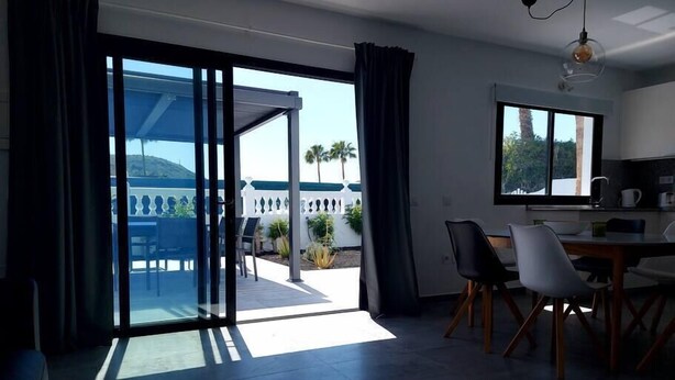 Un salon moderne doté d’une porte vitrée donnant sur une terrasse extérieure, d’un coin repas avec des chaises et d’un espace cuisine avec une fenêtre.