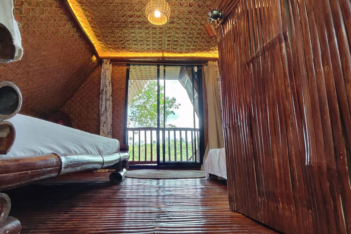 A room with a sloped ceiling, wooden beams, a bed with white bedding, and a view of greenery outside.