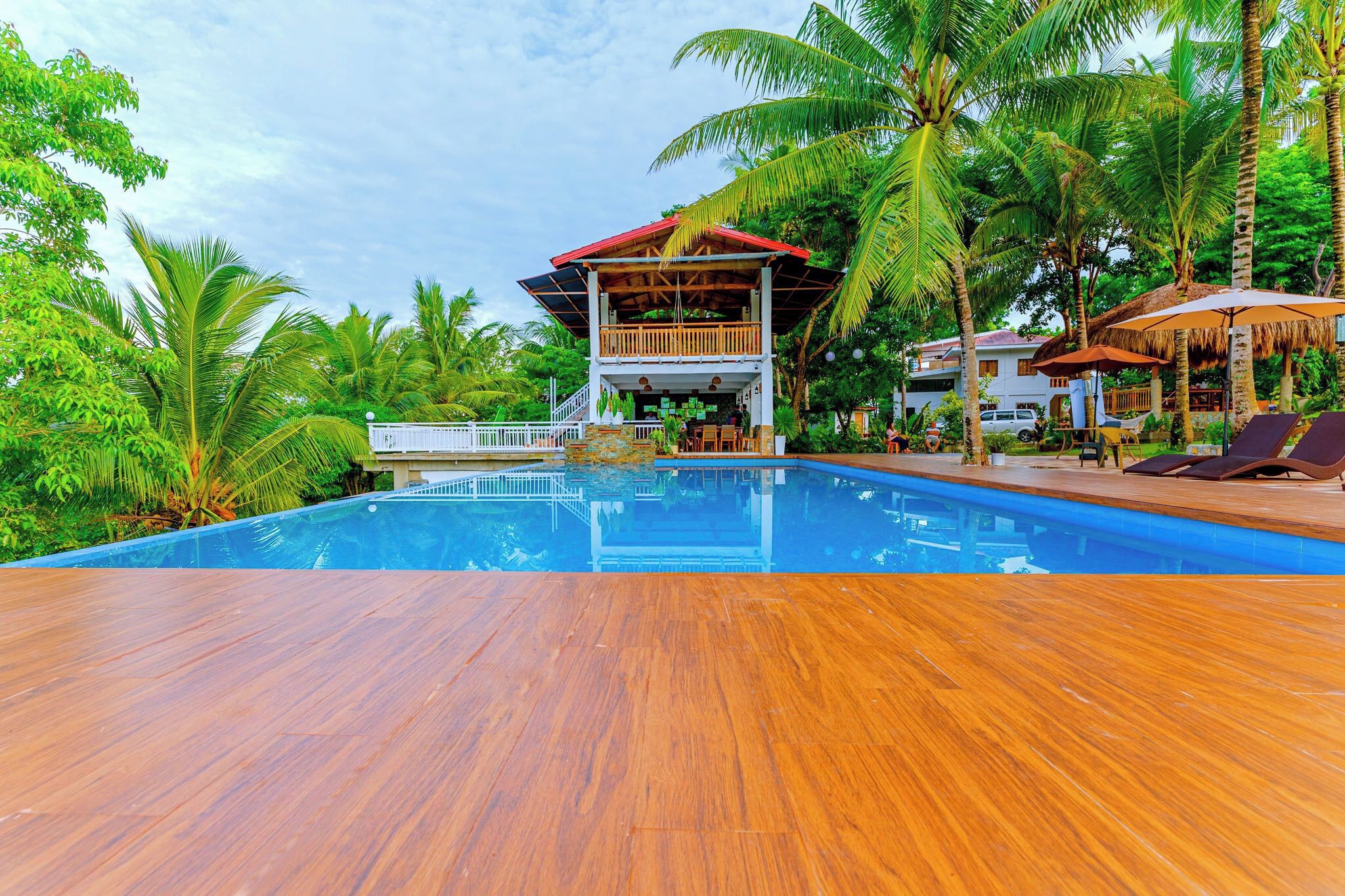 A pool area with wooden decking, a lounge chair, and a building with a red roof and wooden balcony.