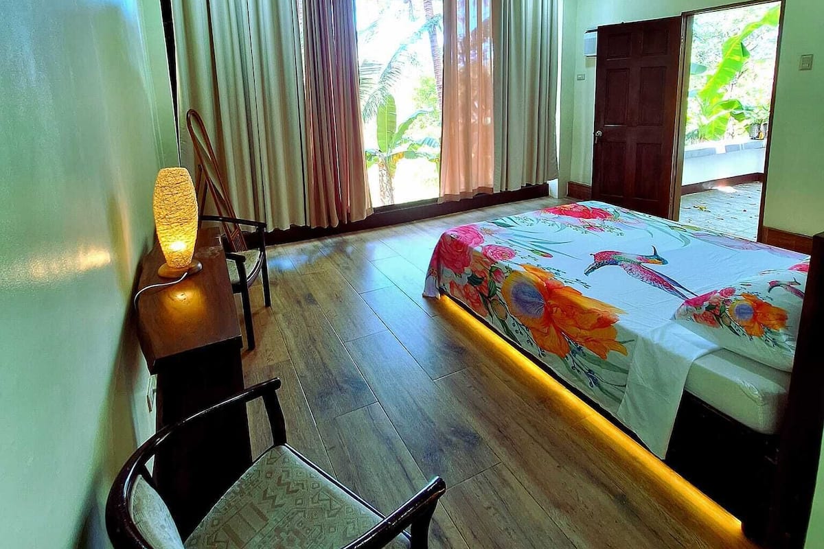 A bedroom with a wooden ceiling, a bed with a floral bedspread, a desk with a lamp, a chair, and a window with curtains.