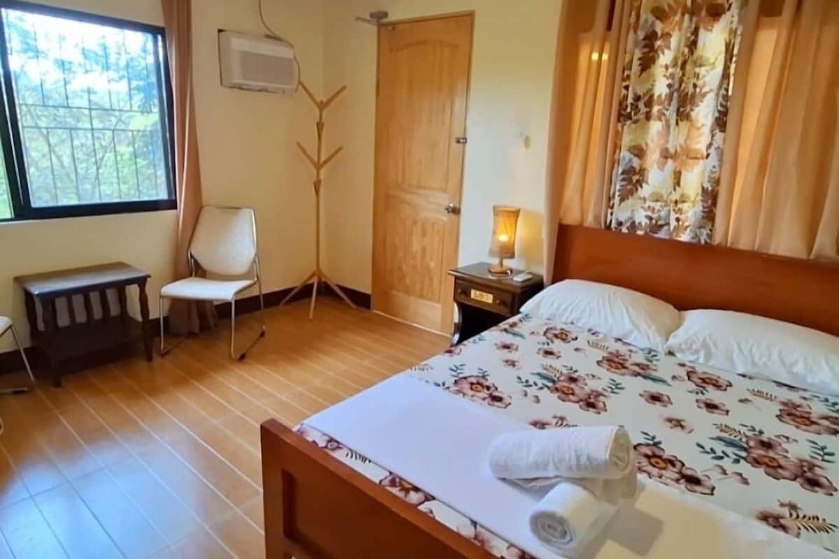 A bedroom with a wooden bed, a bedside table with a lamp, a chair, a bench, and a window with curtains.