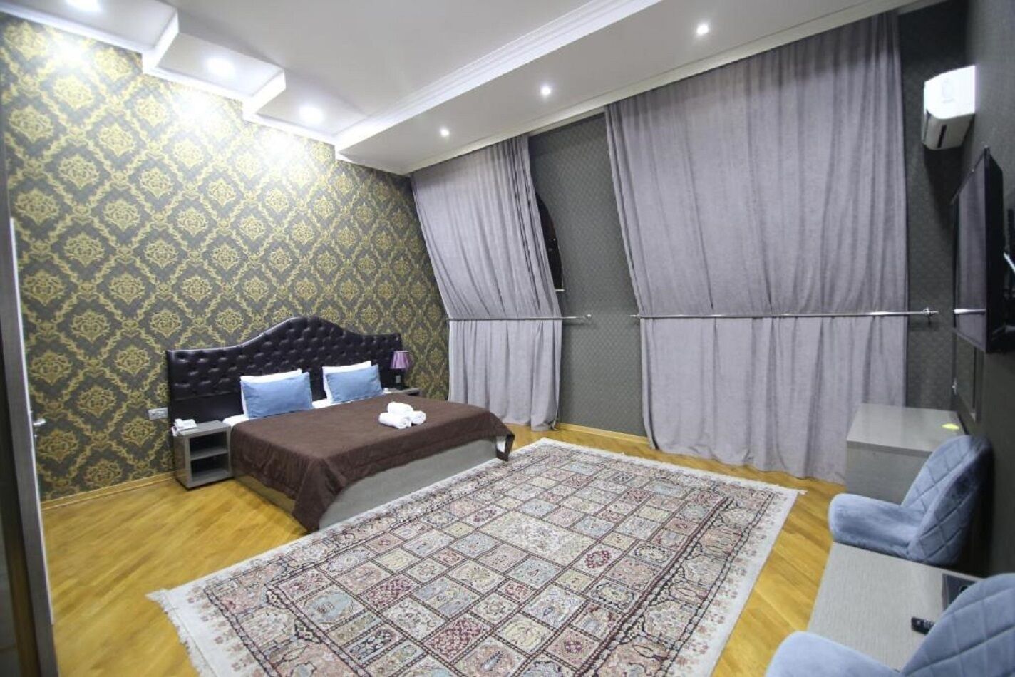 Ein Schlafzimmer mit einem Bett, einem Nachttisch, einem Sessel, einem großen Teppich und Vorhängen.