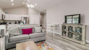 A modern kitchen with white cabinets, a grey sofa with patterned cushions, a wooden coffee table, and a TV on a cabinet. - Sea Glass Paradise Pet-Friendly 5 Mins From Beach! (Pensacola)