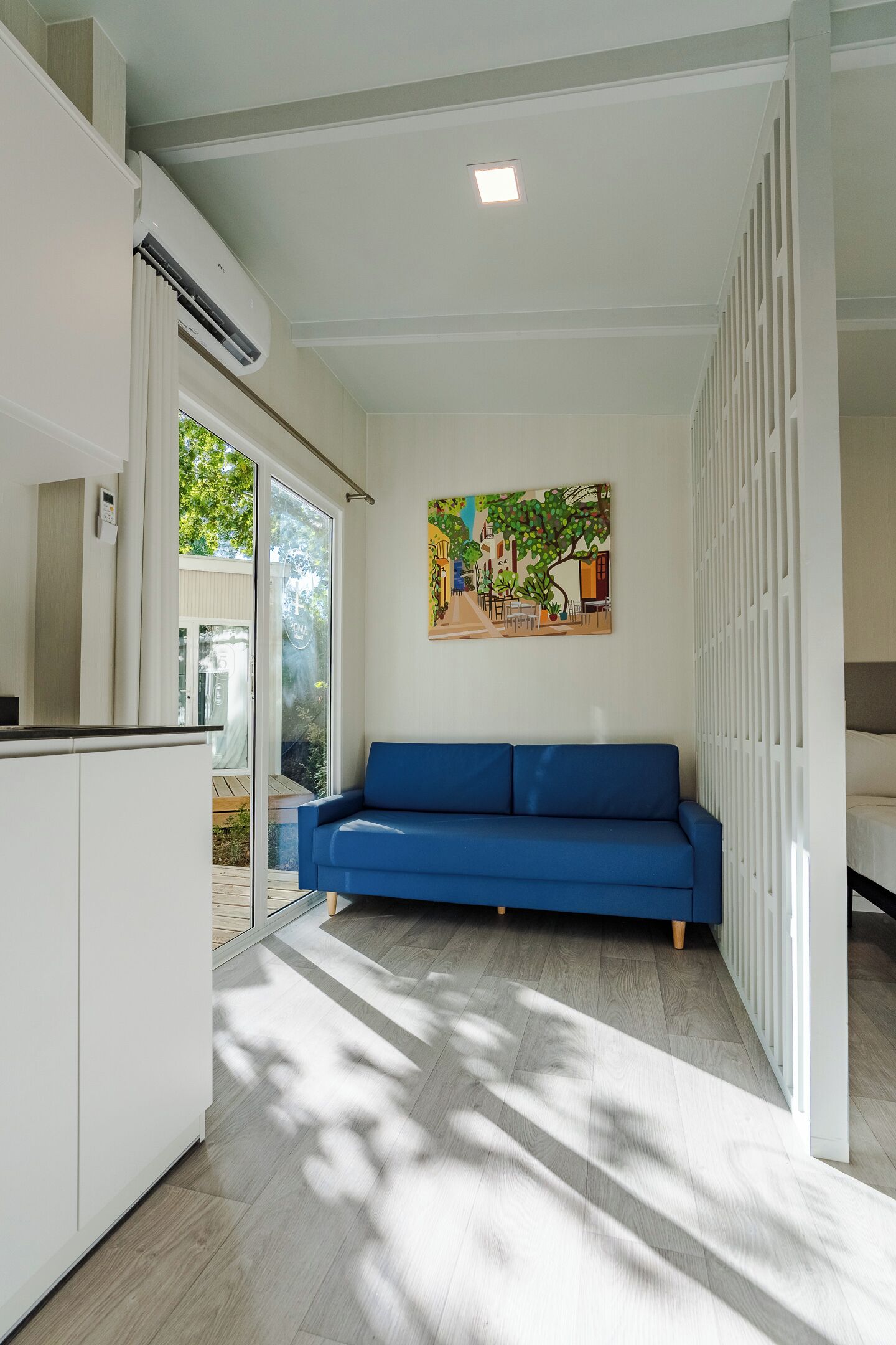 A modern living room with a blue sofa, a painting on the wall, and a sliding glass door leading to an outdoor area.