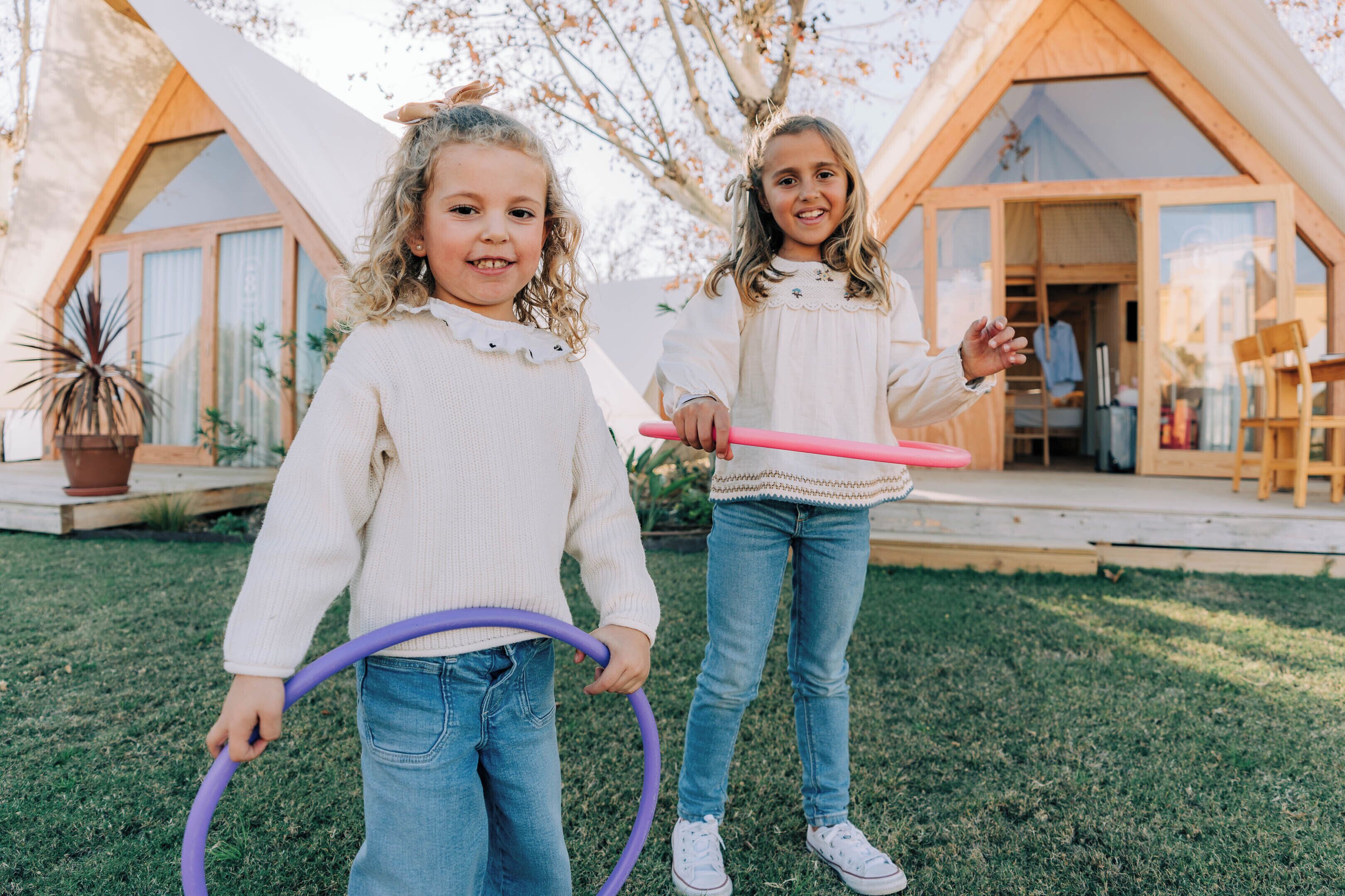 Two children playing with hula hoops in a grassy area in front of a wooden cabin with large windows and a door.