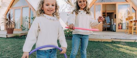 Two children playing with hula hoops in a grassy area in front of a wooden cabin with large windows and a door.