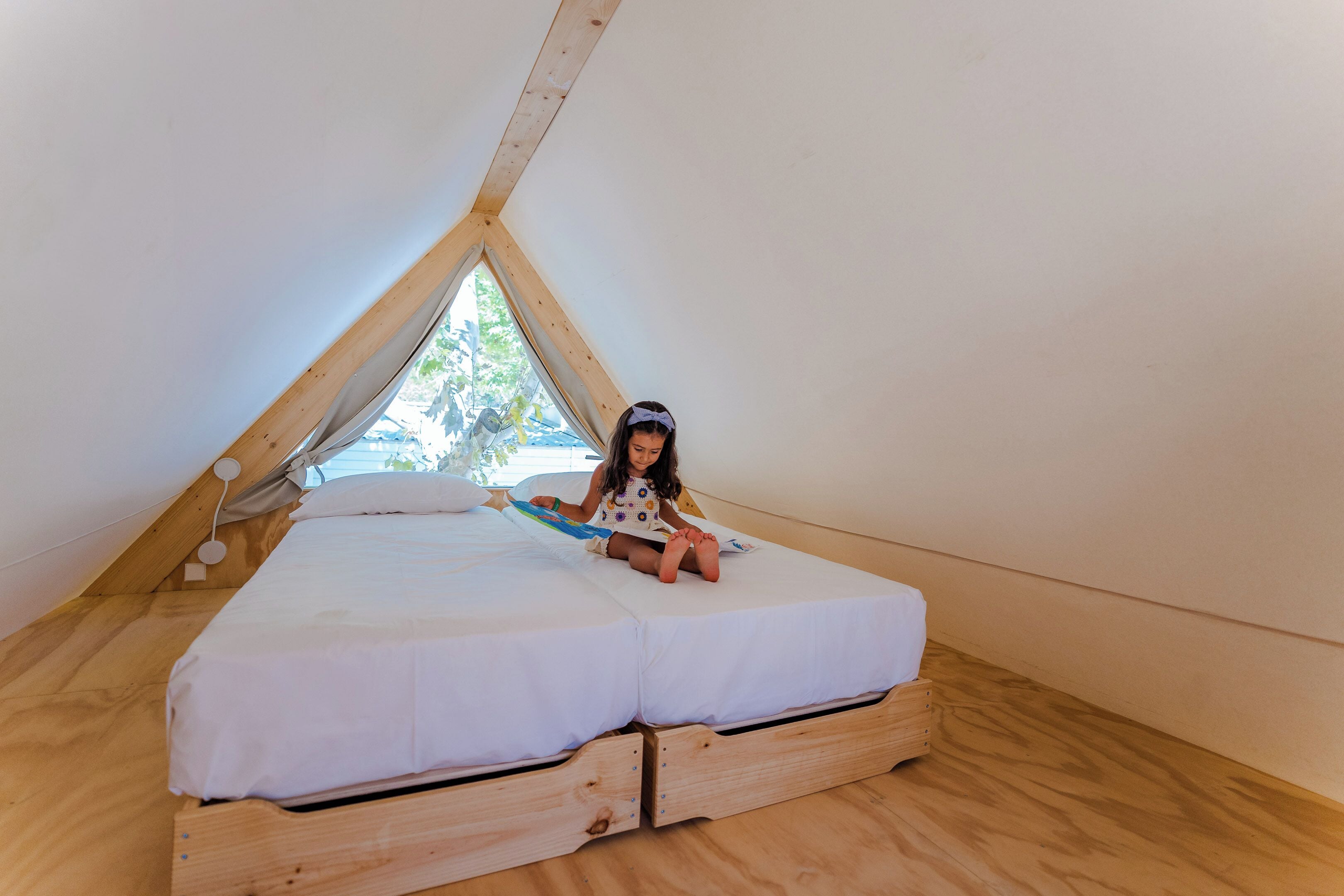 A child sitting on a wooden bed in a room with a sloped ceiling and a window.