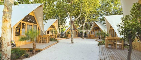 A series of triangular tents with wooden decks and seating areas, surrounded by trees and a sandy ground.