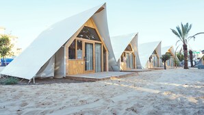 Una hilera de cabañas de playa con techos triangulares y exteriores de madera, ubicada sobre suelo arenoso con palmeras al fondo.