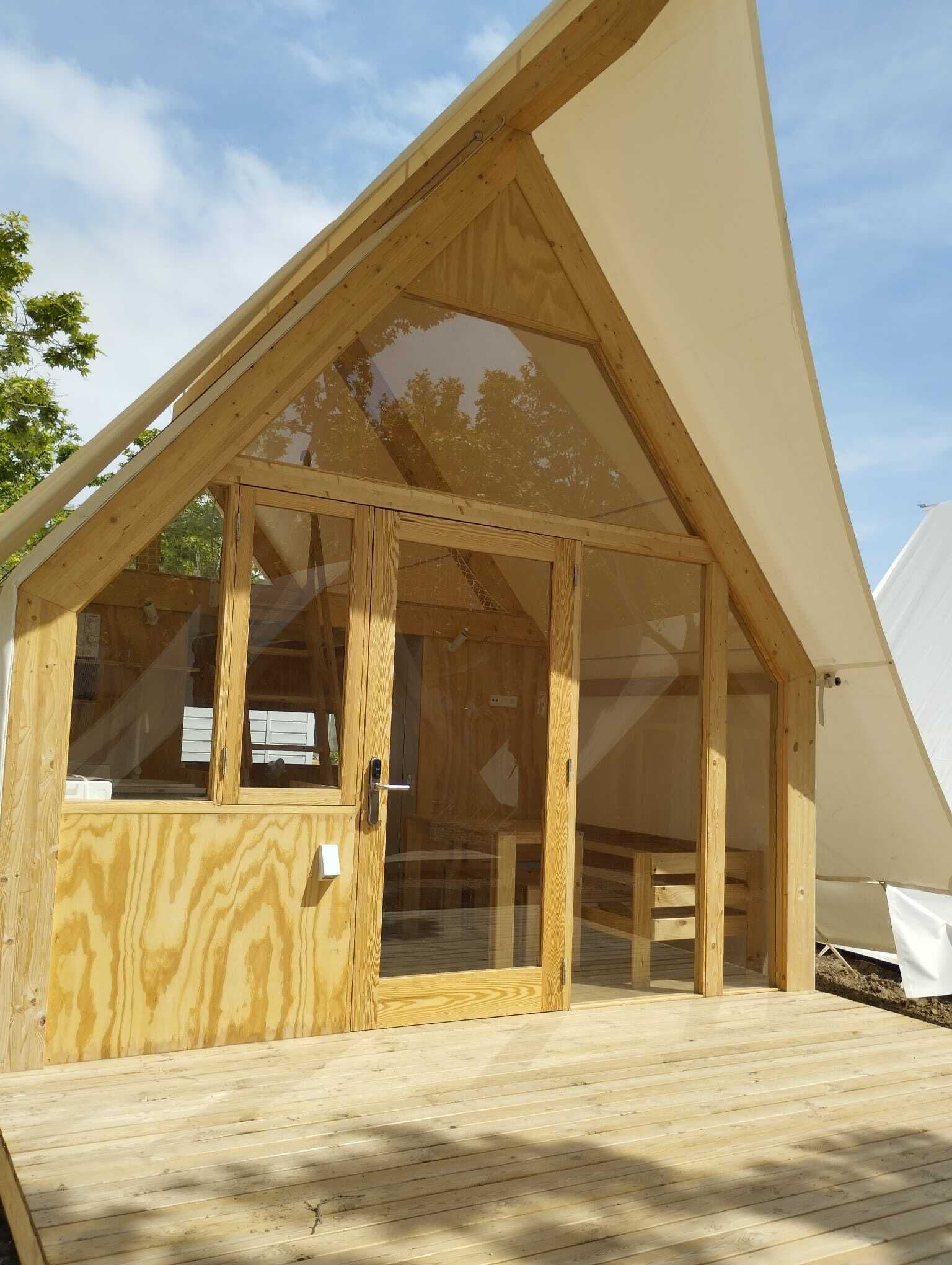 Il s’agit d’une structure en bois dotée d’un toit à deux versants et de grandes portes vitrées, qui semble être un espace d’hébergement temporaire ou un lieu destiné à accueillir des événements.