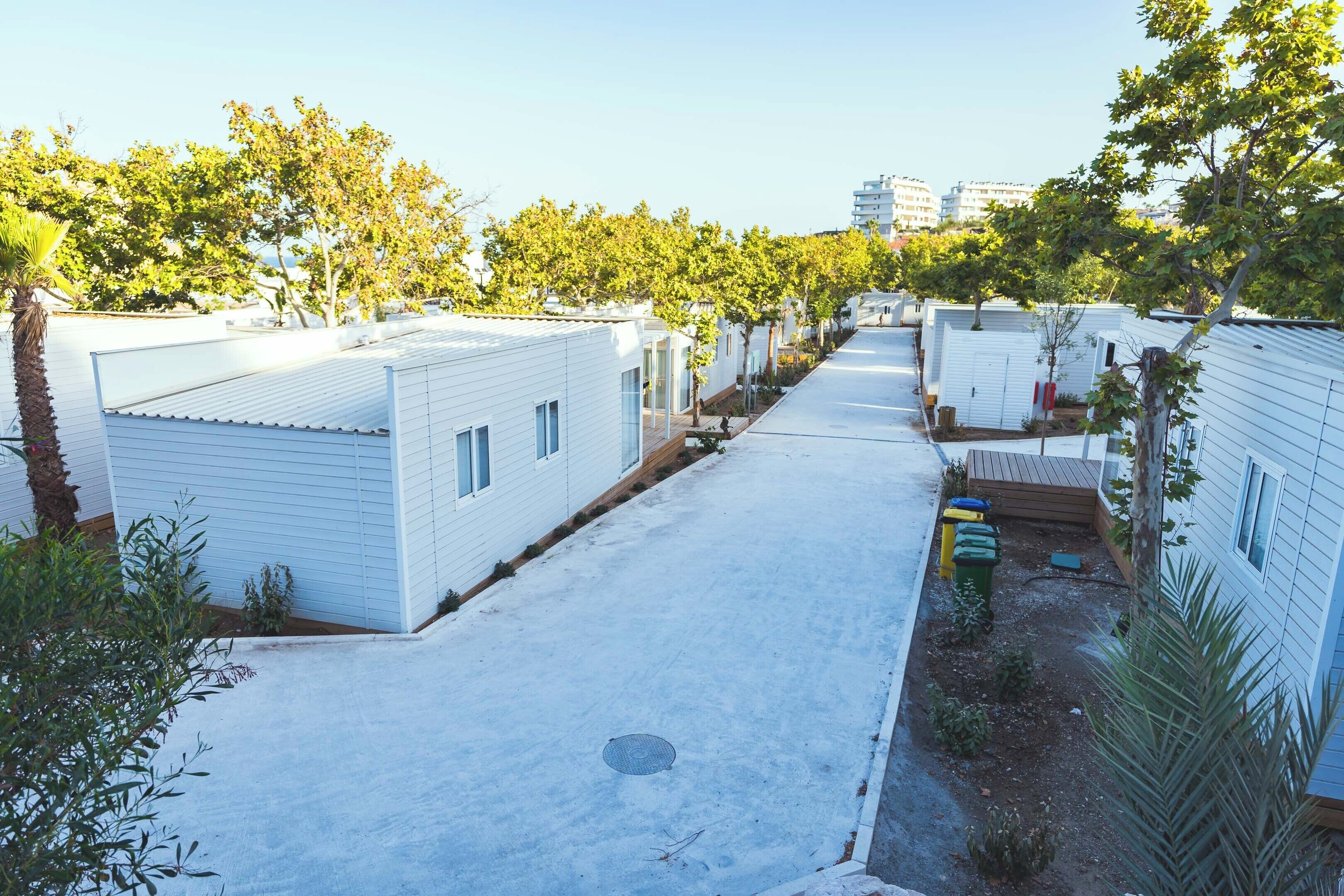 Una hilera de viviendas modulares blancas con un camino pavimentado y árboles al fondo.
