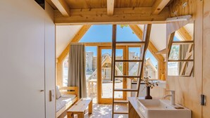 Una habitación con techo de madera, un ventanal grande, una cama, un banco, un lavamanos y un espejo.