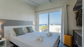 Uma cama bem arrumada com lençóis e almofadas brancas, uma mesa de cabeceira com abajur, uma mala e uma varanda com vista.