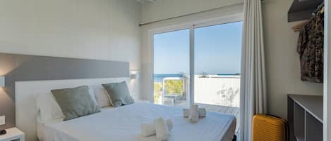 A neatly made bed with white linens and pillows, a bedside table with a lamp, a suitcase, and a balcony with a view.