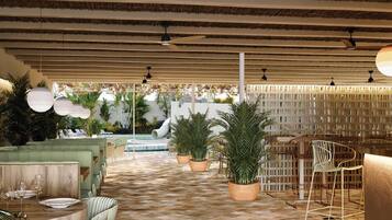 Un área de comedor al aire libre con suelo a cuadros, mesas redondas y sillas. Hay plantas en macetas y una piscina al fondo.