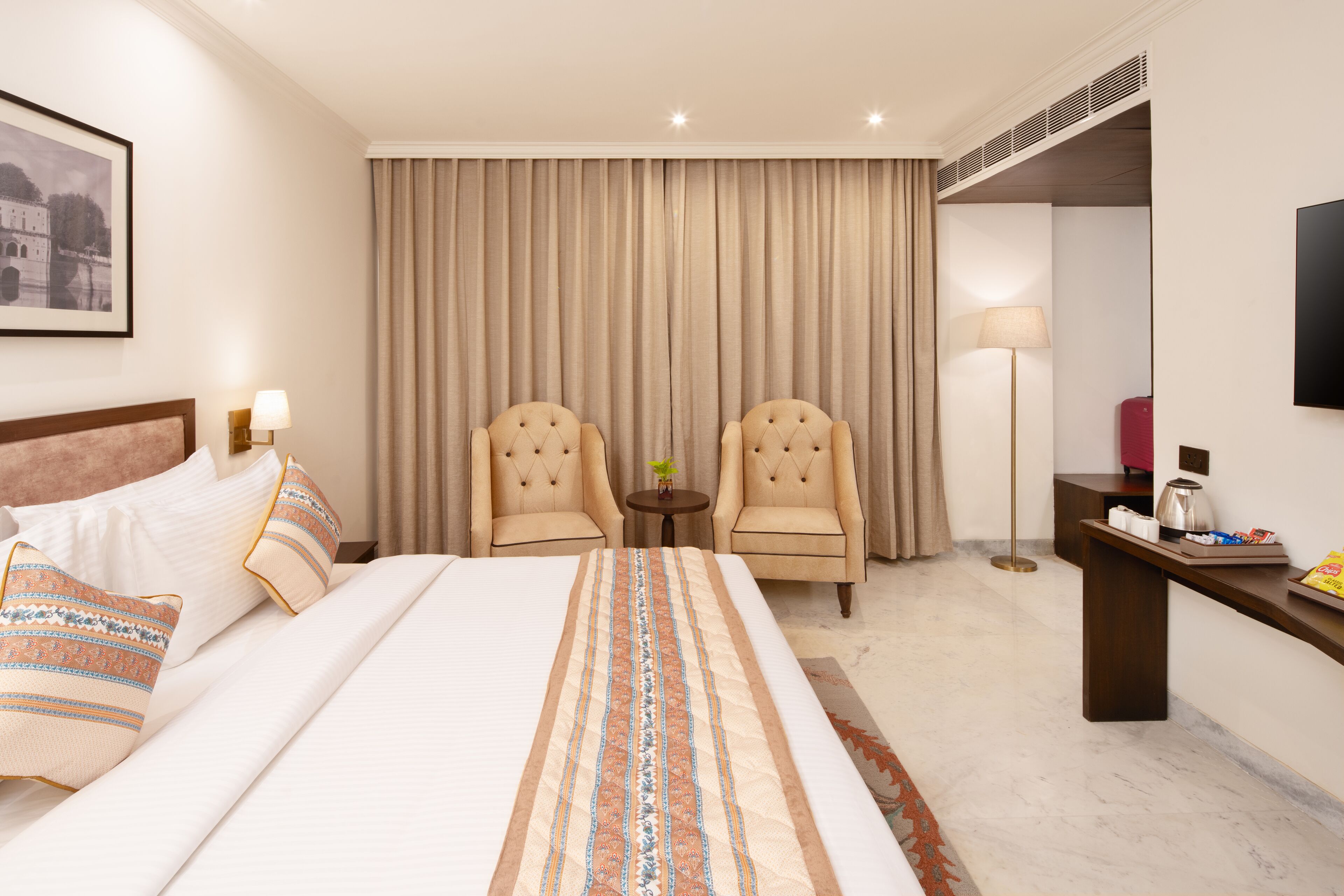 A hotel room with a large bed, two armchairs, a small table, a desk, and a television.