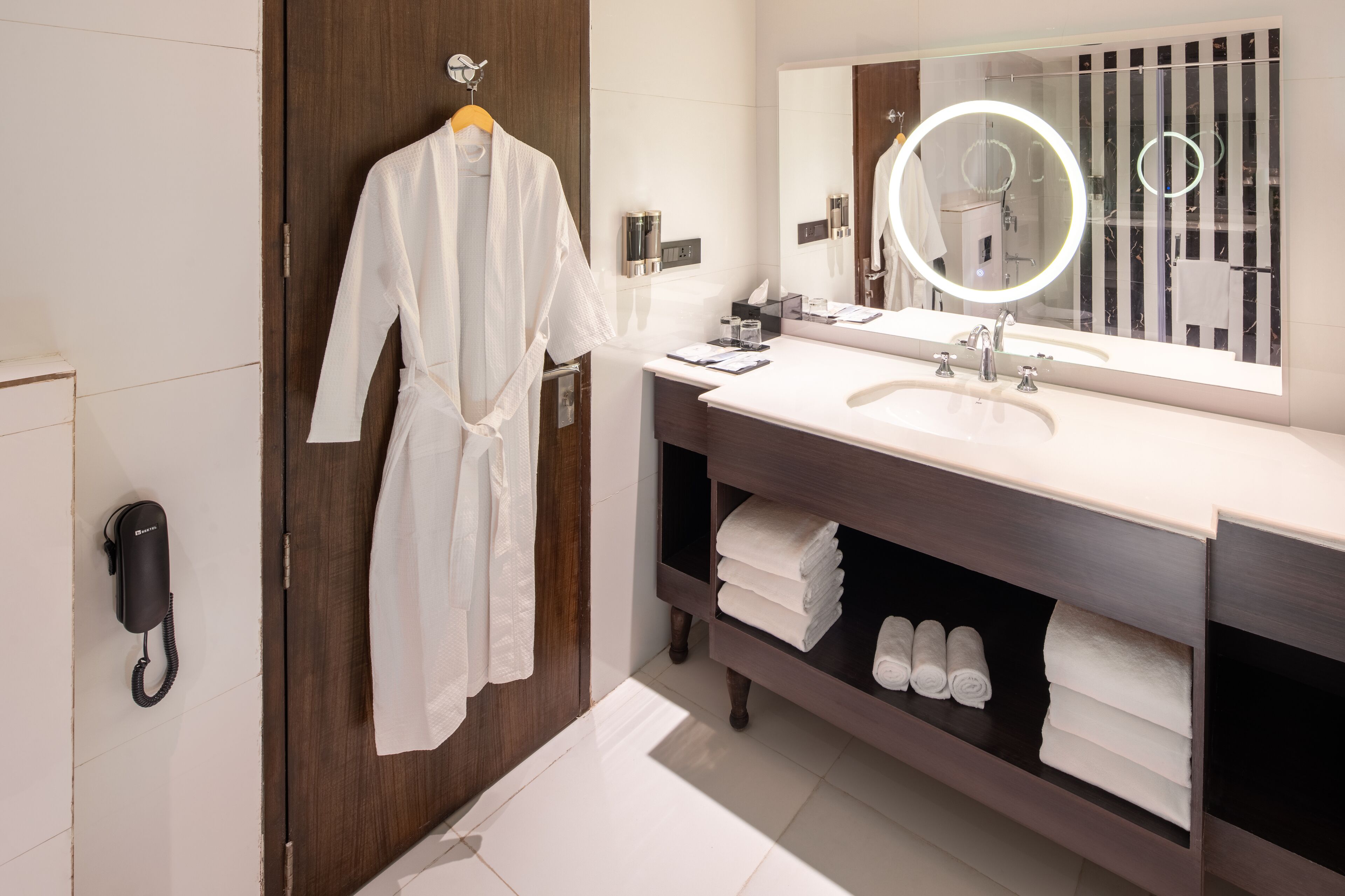 A modern bathroom with a wooden vanity, a large mirror with a circular light, and a robe hanging on a rack.