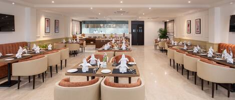 A spacious hotel dining area with multiple tables and chairs, a central bar, and framed artwork on the walls.