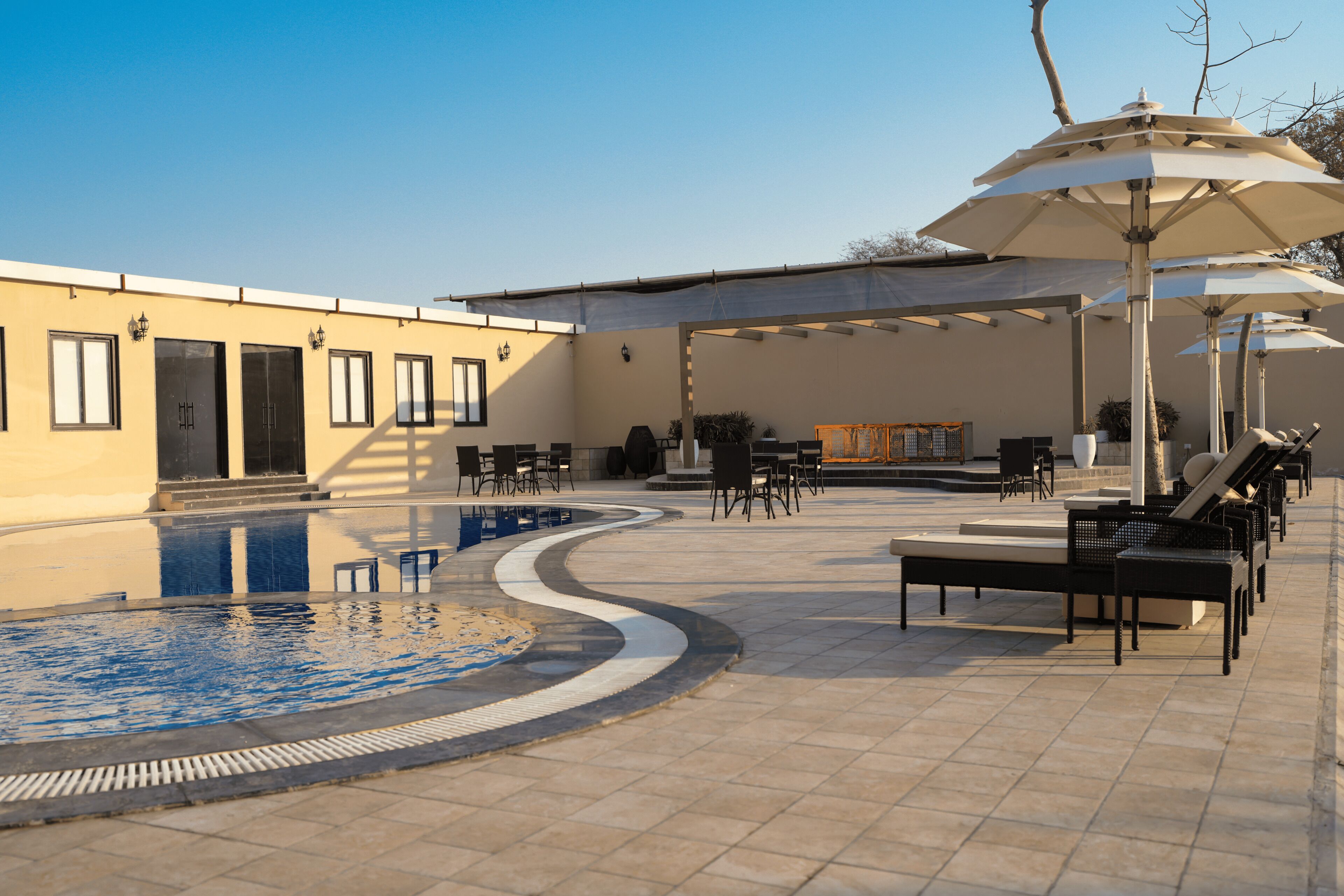 A pool area with lounge chairs and umbrellas, a covered seating area, and a building with doors and windows.