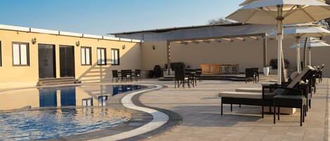 A pool area with lounge chairs and umbrellas, a covered seating area, and a building with doors and windows.