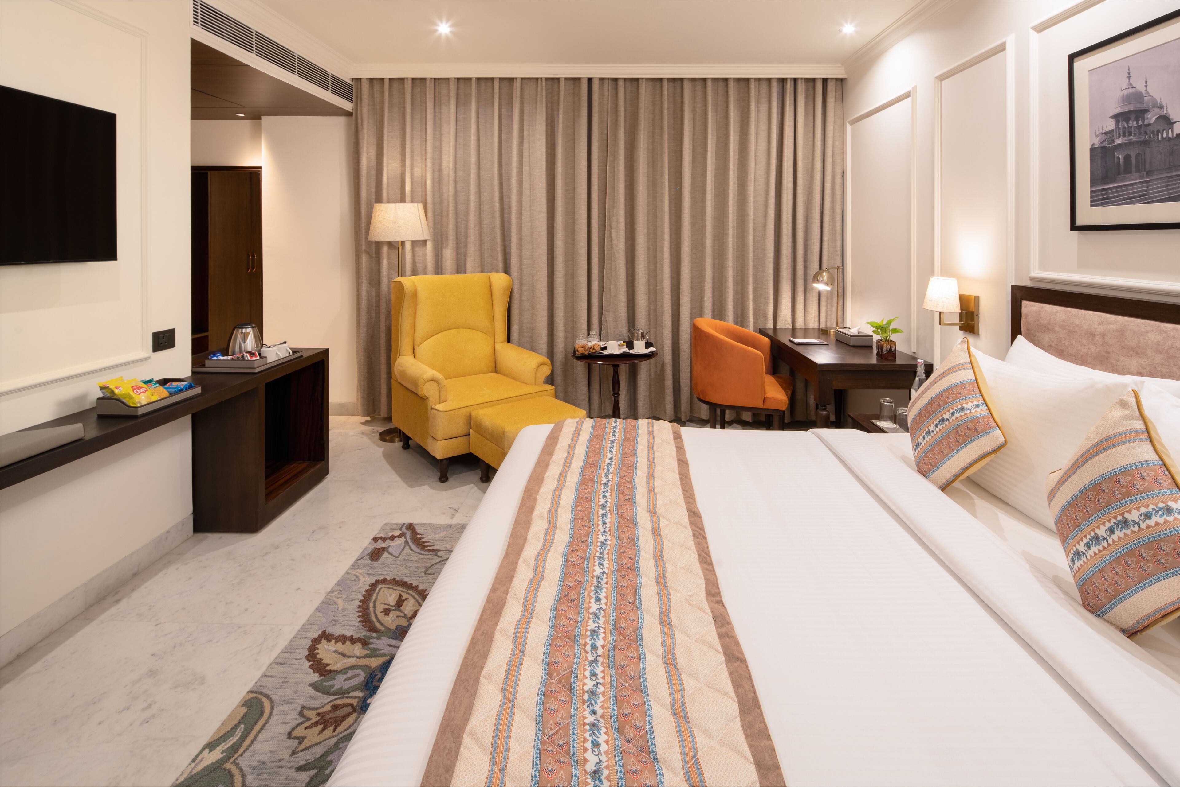 A hotel room with a large bed, a desk, a yellow armchair, an orange chair, a small table, and a TV mounted on the wall.