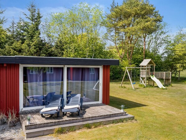 Une terrasse extérieure moderne avec deux chaises longues, un bâtiment rouge doté de portes vitrées et une structure de jeu en bois avec un toboggan.