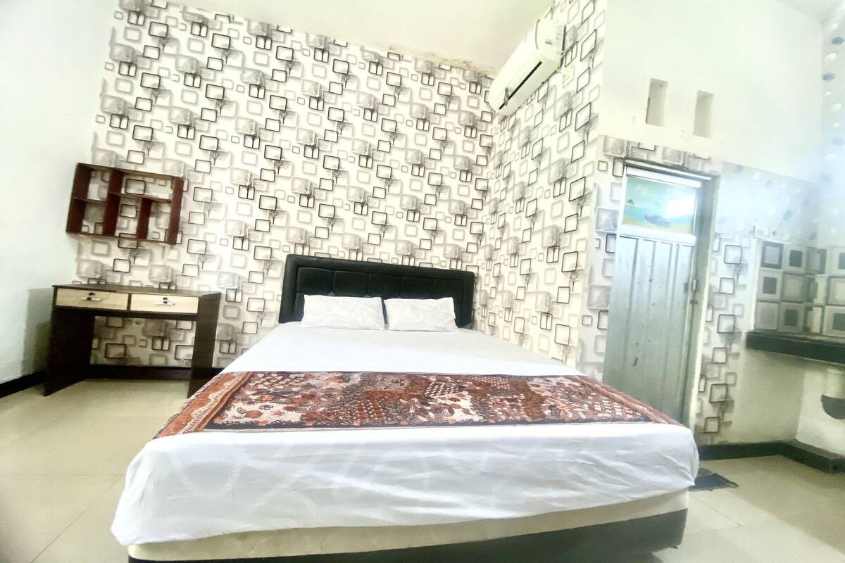 A hotel room with a patterned wall, a bed with a white and brown bedspread, a nightstand with a mirror, and a wardrobe.