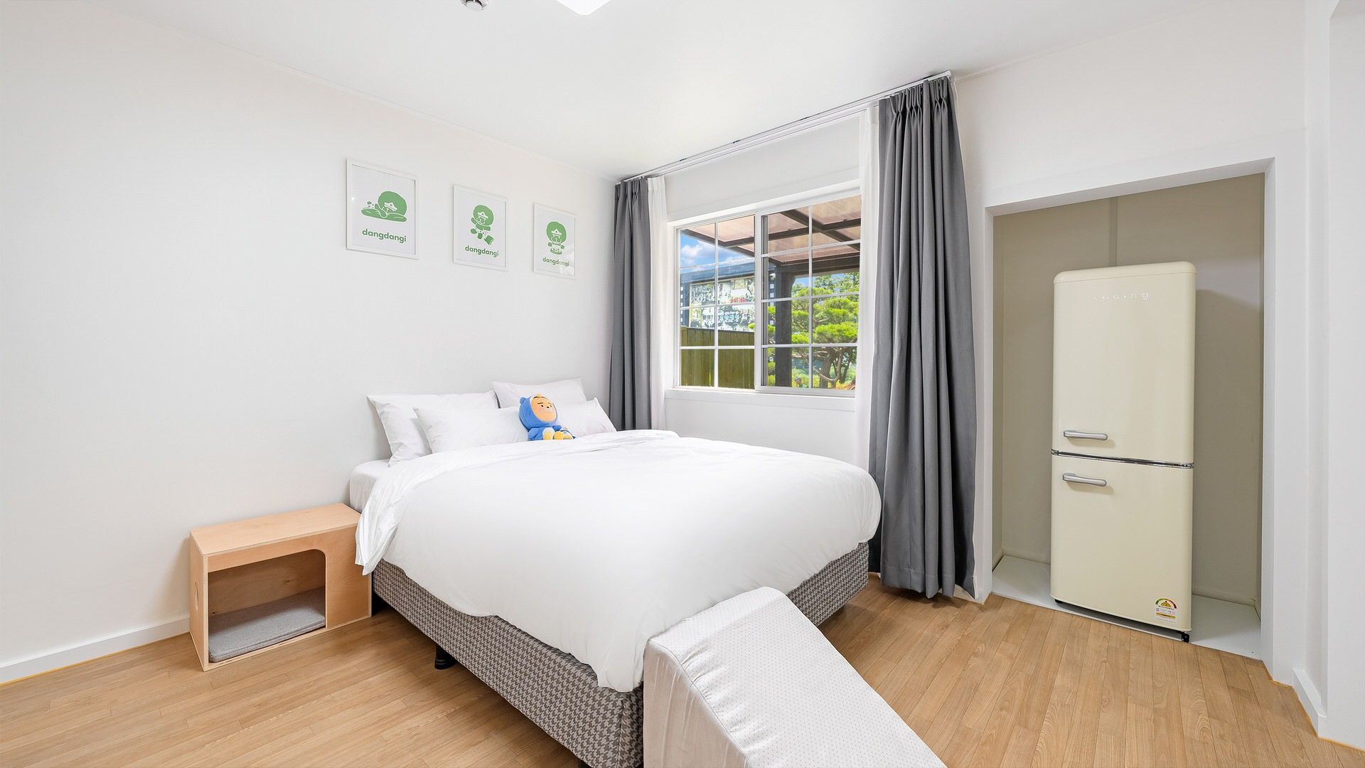 A bedroom with a bed, a nightstand, a window with curtains, and a refrigerator.