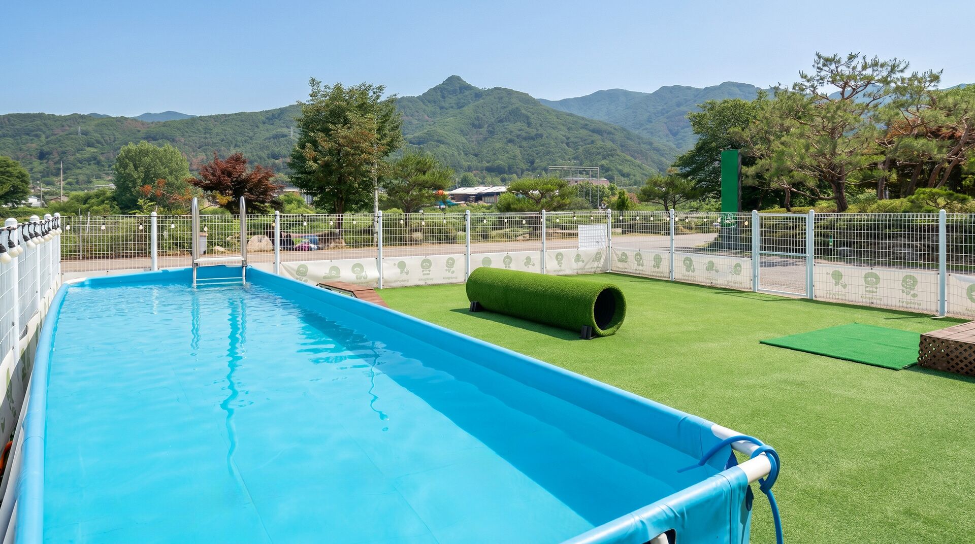 Una piscina con una zona de césped artificial verde, rodeada por una valla blanca, y montañas al fondo.