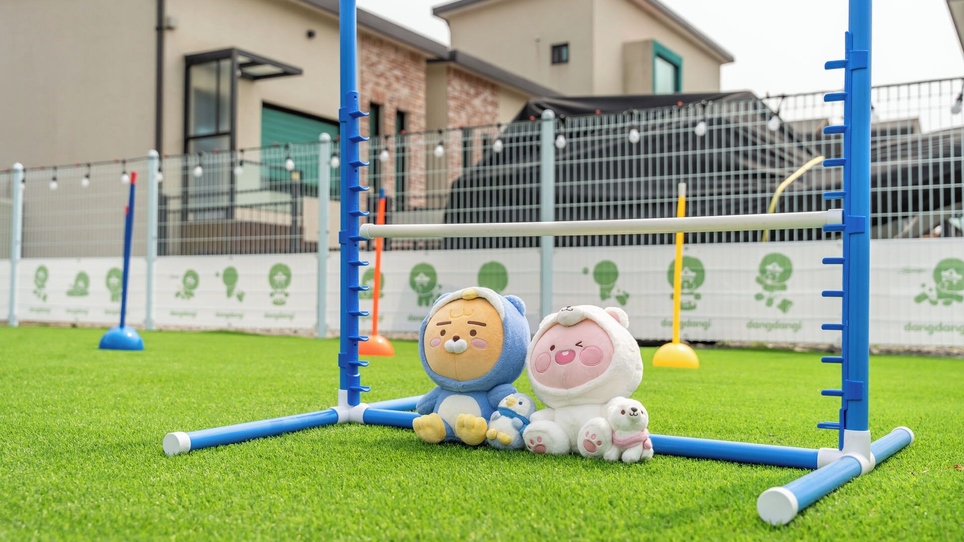 Un'area giochi per bambini con una struttura colorata in blu e bianco, un prato sintetico verde e una recinzione con un logo.