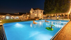 Ein Schwimmbecken mit Luftmatratzen, eine Holzterrasse und ein Gebäude mit zahlreichen Fenstern und einer Veranda.