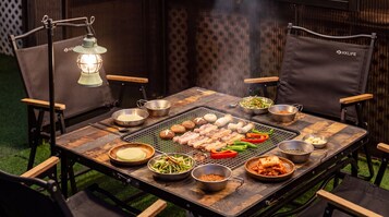 A portable grill with various meats and vegetables cooking, surrounded by chairs and dining tables.