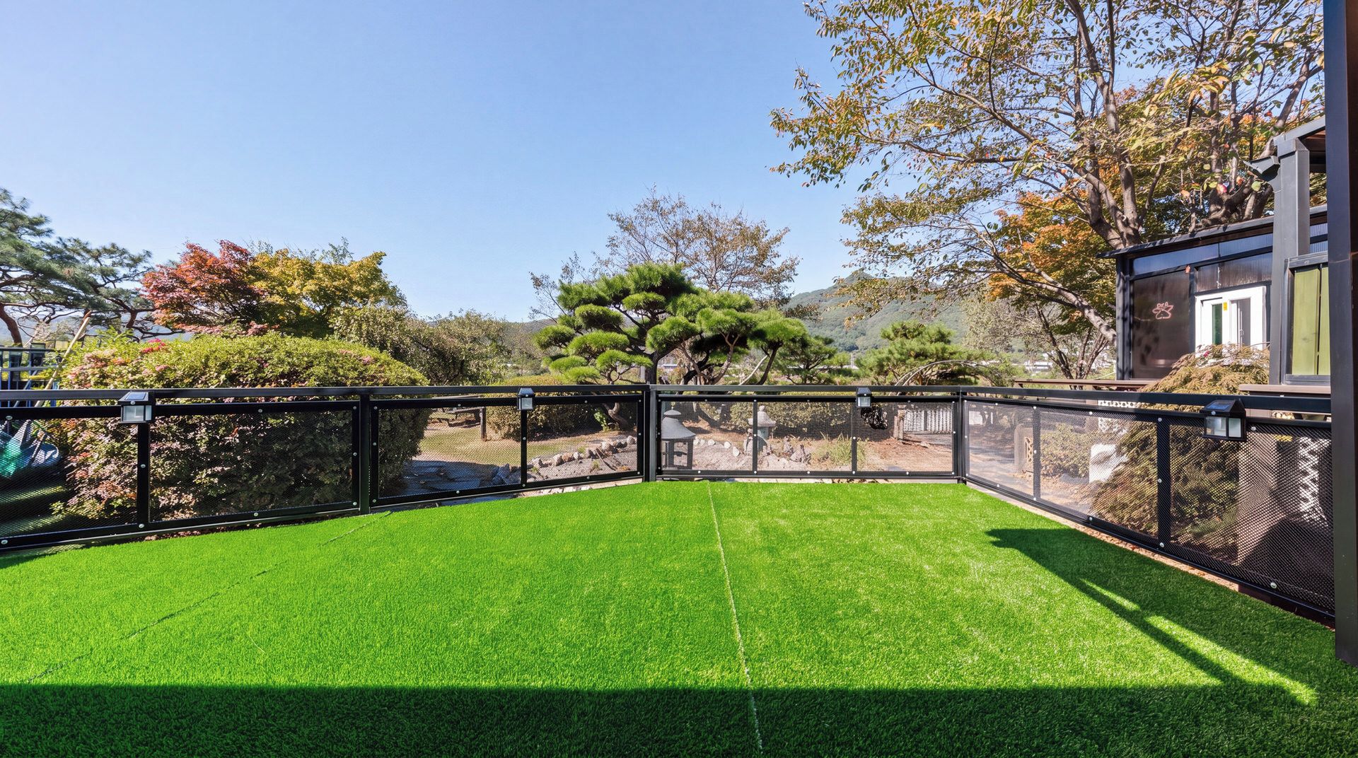 A fenced outdoor area with artificial grass, a tree, and a building in the background.